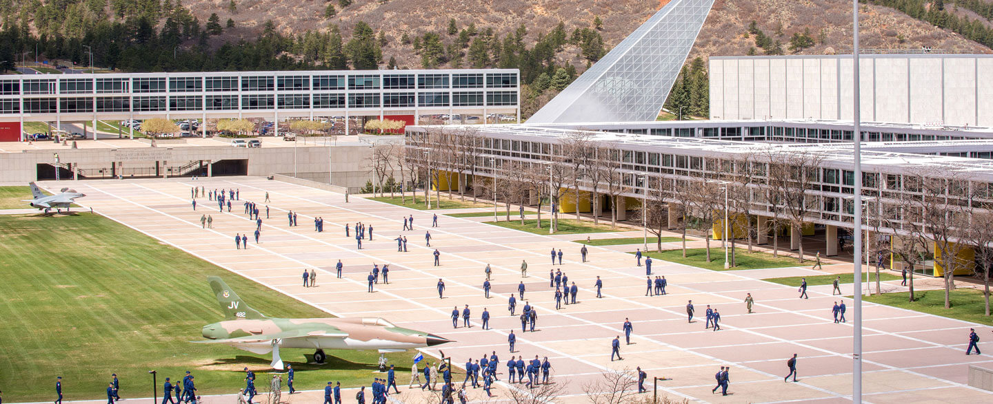 Cadets walking across terrazzo near F-16 display with chapel spires and mountains visible