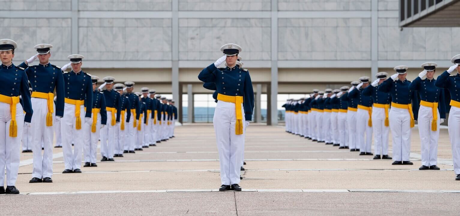 Cadets in blue dress uniforms with gold sashes saluting in formation on parade ground