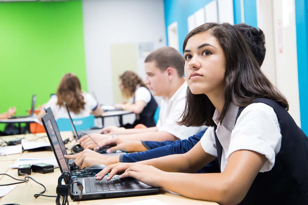 Students in navy vests and white shirts focused on laptops in vibrant green and blue classroom