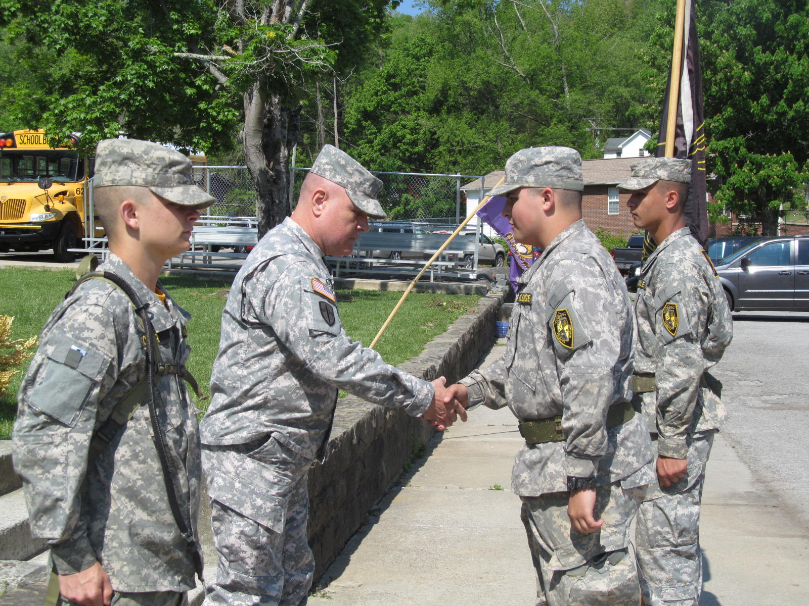 Cadets in ACU uniforms during outdoor formation with staff member, school buses in background
