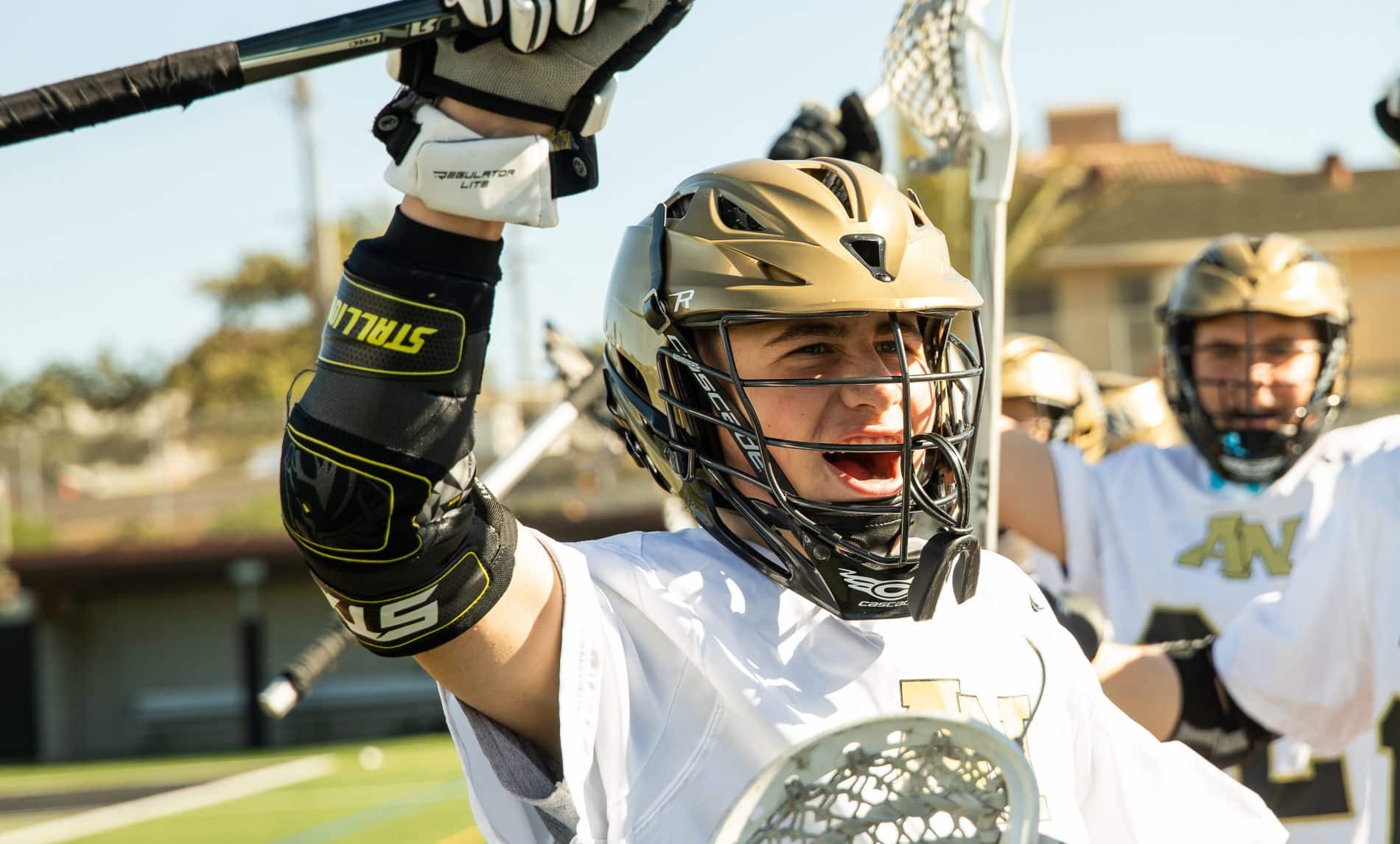 Cadet lacrosse player in gold helmet and white jersey celebrating with stick raised, teammates behind