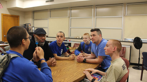 Cadets in blue shirts gathered around table in classroom during group discussion with instructor