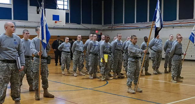 Cadets in gray PT shirts and camo pants standing in formation in gymnasium holding unit guidons