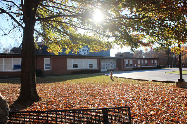 Red brick academy building with BCA crest on facade, autumn leaves on lawn, sunlight through trees