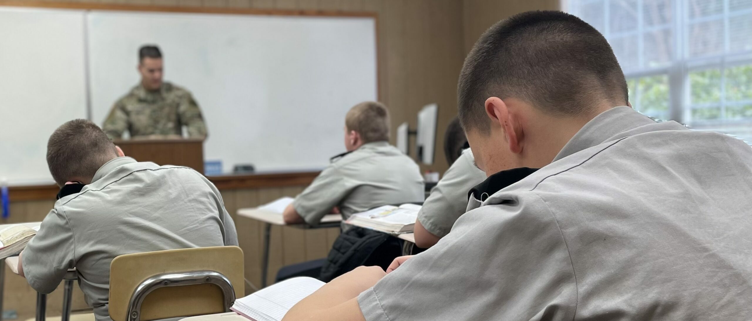 Cadets in gray uniforms studying at desks while instructor in camouflage teaches at whiteboard