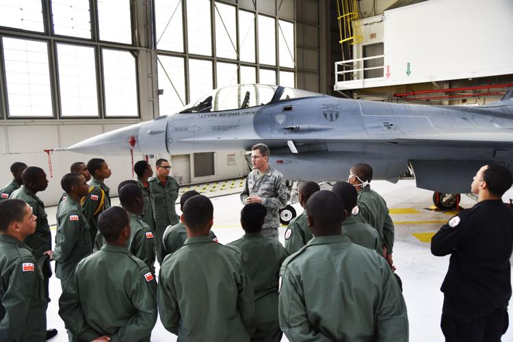 Cadets in olive flight suits receiving briefing from Air National Guard instructor next to F-16 fighter jet in hangar