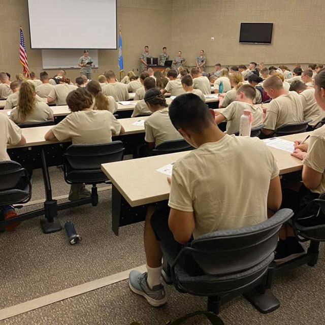 Cadets in tan PT shirts attending lecture in tiered classroom with military instructors