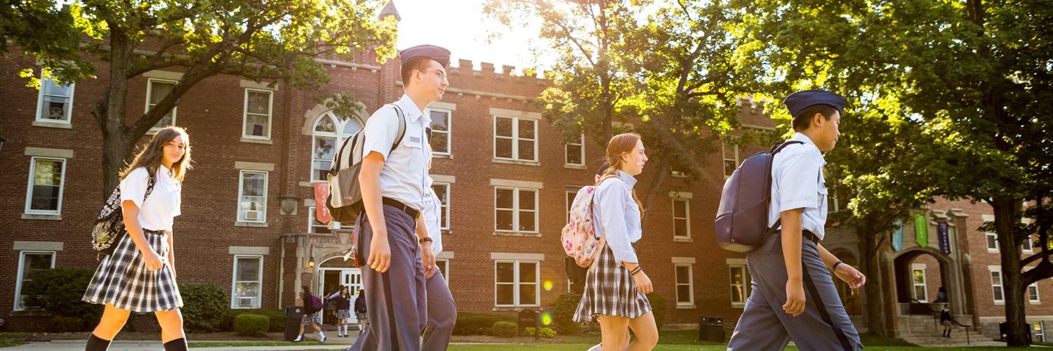 Students walking on campus with cadets in gray uniforms and girls in plaid skirts near historic brick building