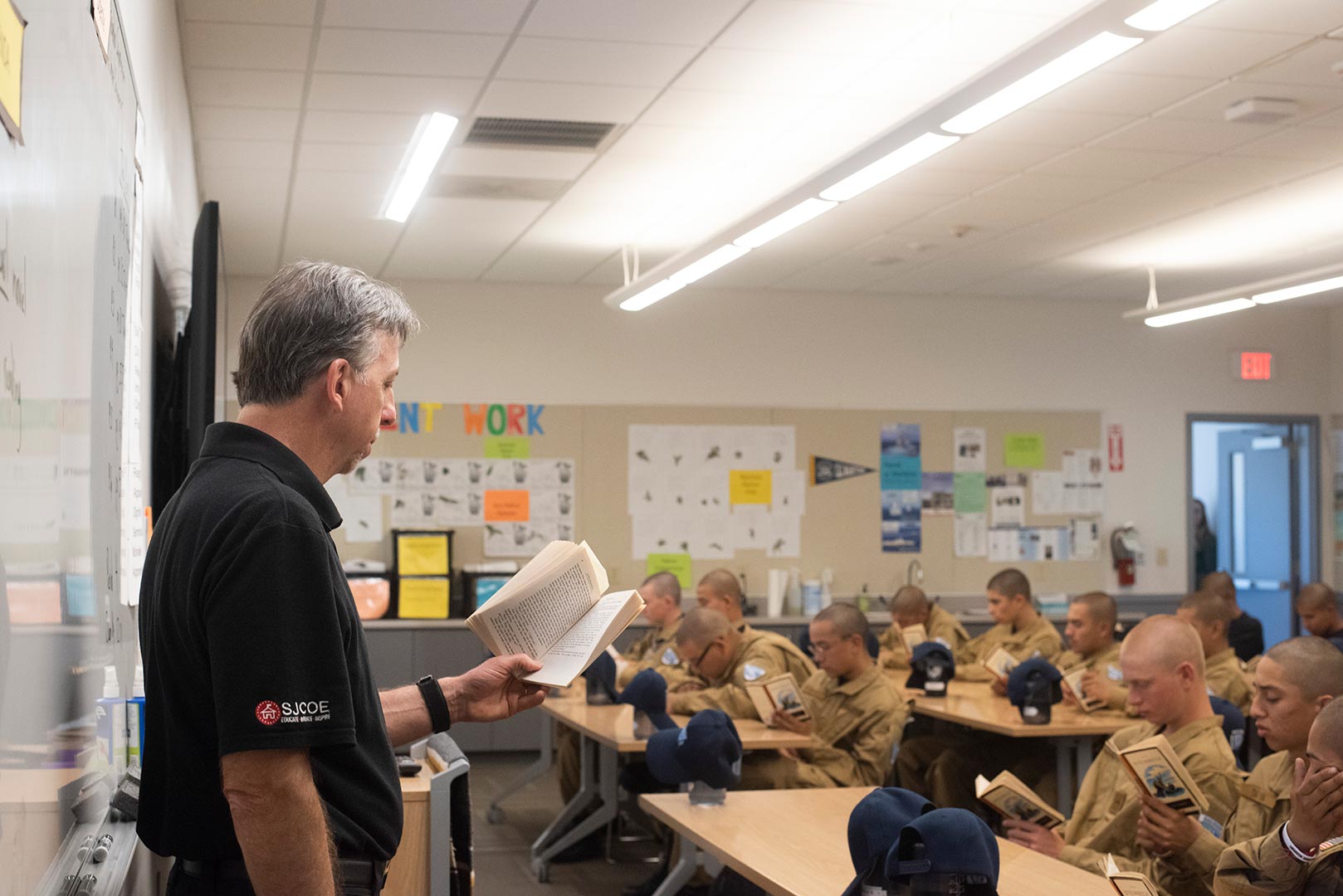 SJCOE instructor reads aloud to cadets with shaved heads in tan uniforms during classroom lesson
