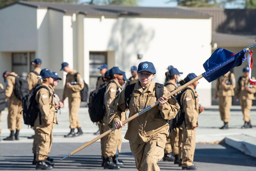 Female cadet carrying platoon guidon flag leads formation in tan uniforms across campus grounds