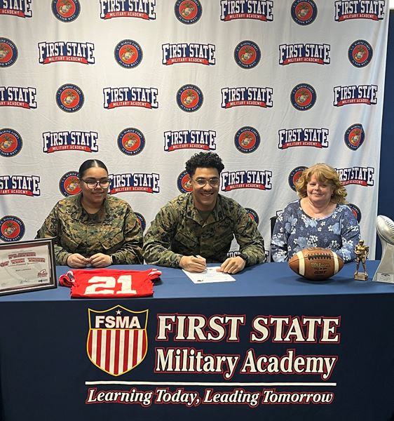 Cadet in camo uniform at college signing ceremony with football, jersey, and trophy at branded table