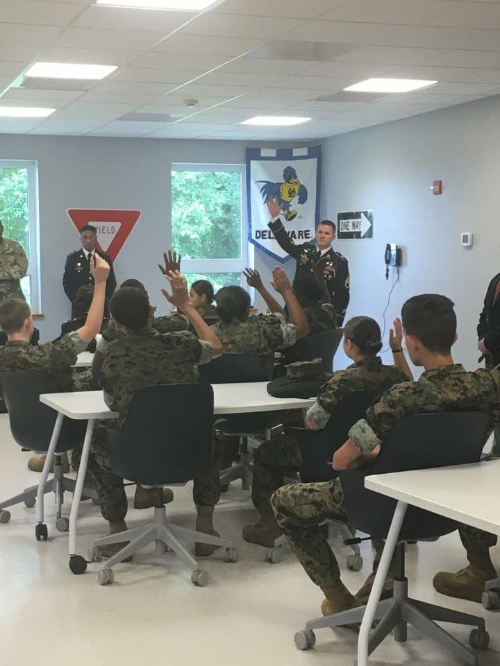 Cadets in Marine camouflage uniforms raising hands during classroom instruction with Delaware banner