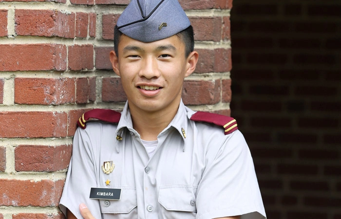 Smiling cadet in gray Class B uniform with garrison cap standing by brick wall