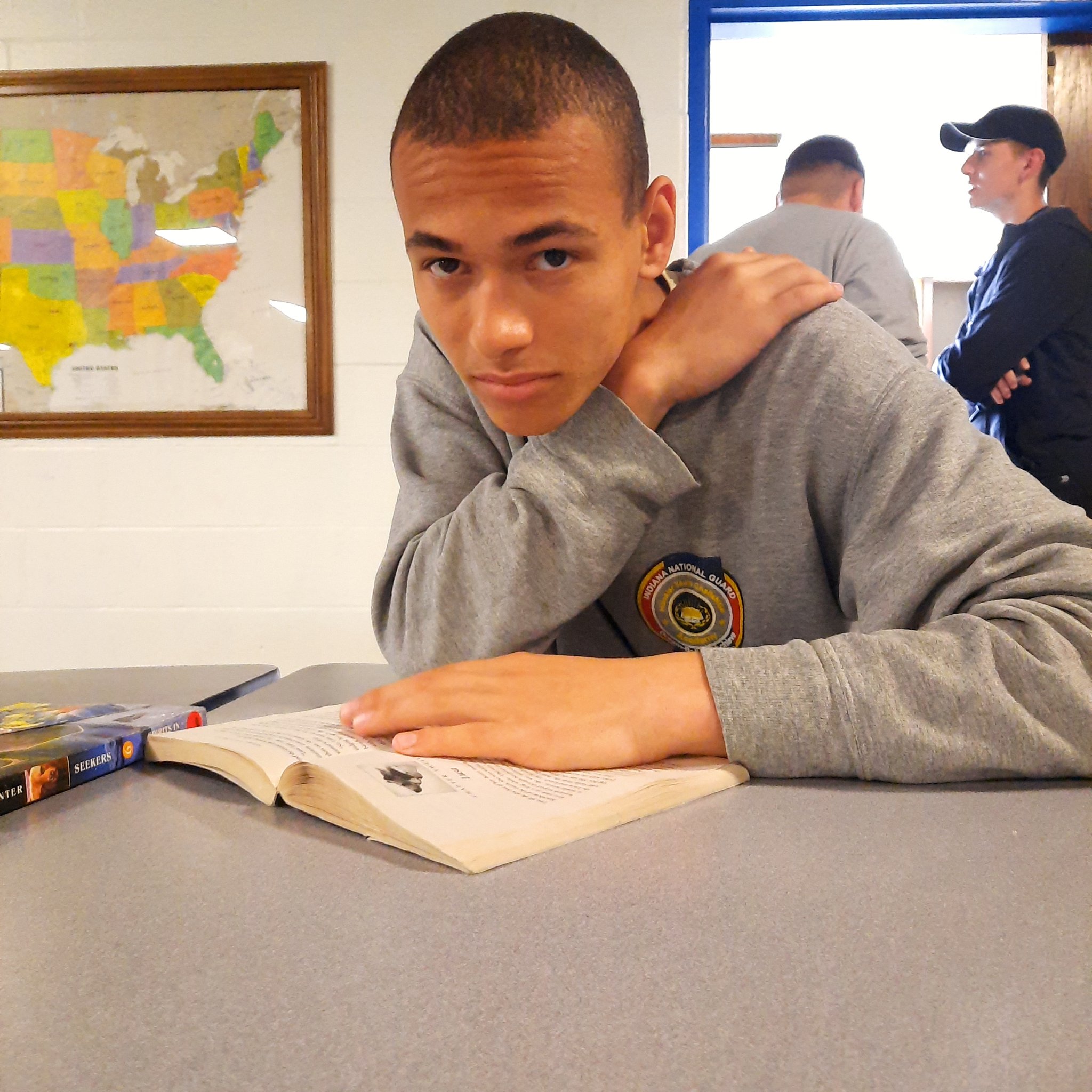 Cadet in gray sweatshirt studying with open book at classroom desk with US map on wall