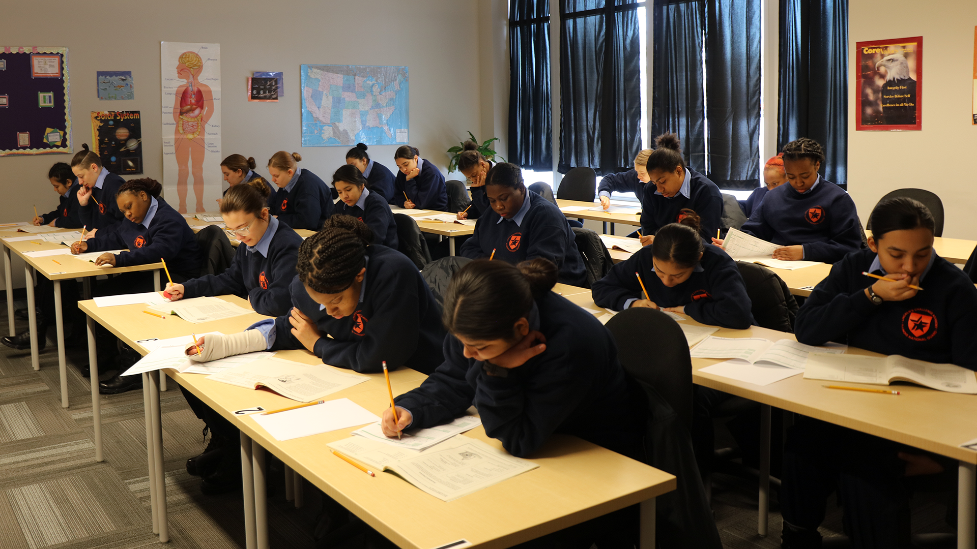 Cadets in navy blue uniforms with patches taking written exam at desks in classroom