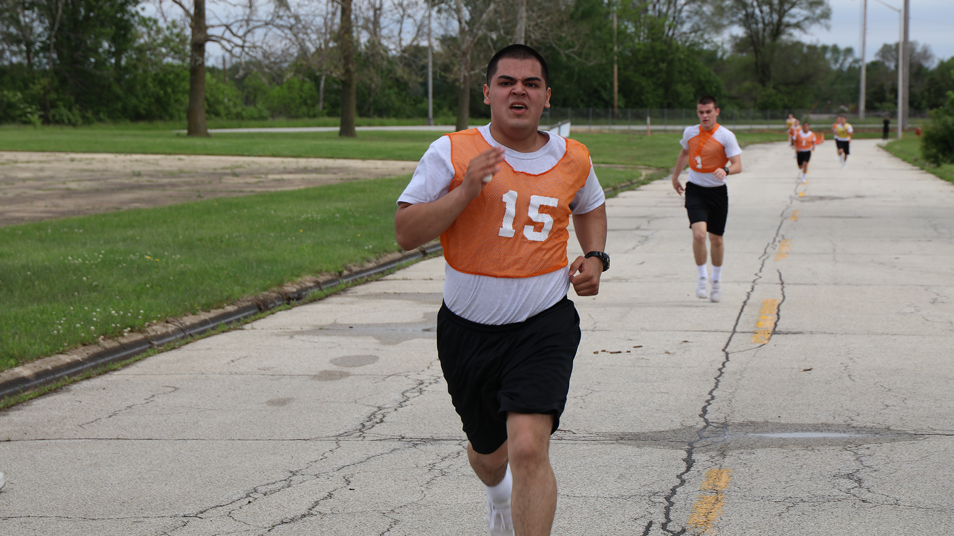 Cadets in orange numbered jerseys running on paved road during cardio training with grass fields nearby