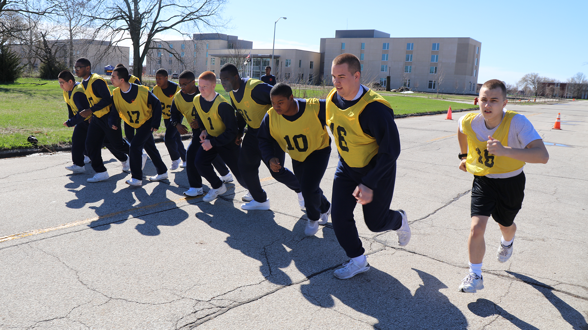 Cadets in yellow jerseys doing high-knee exercises outdoors with campus buildings in background