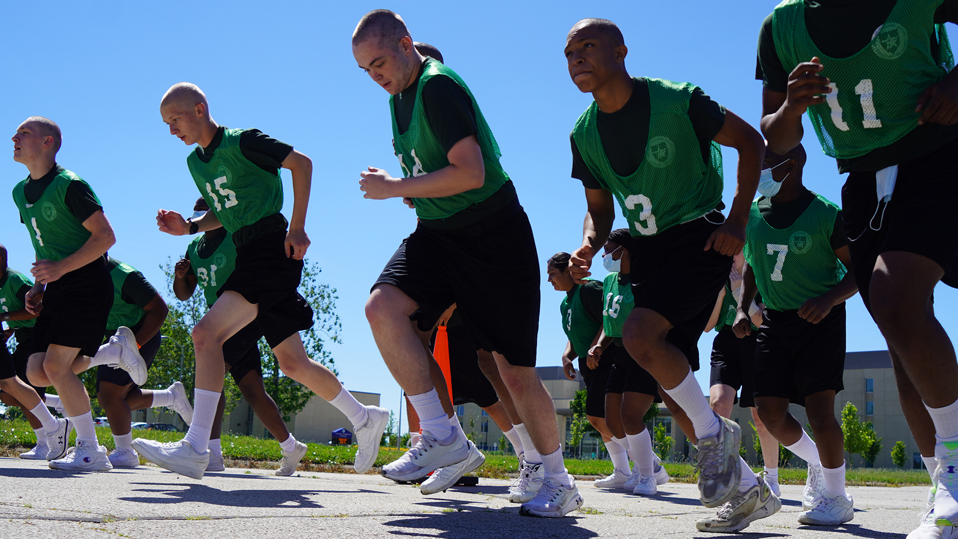 Cadets in green numbered jerseys running in formation during outdoor physical training on sunny day