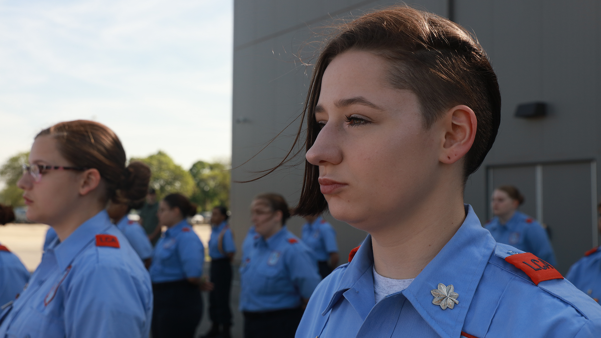 Female cadets in light blue uniforms with ponytails standing at attention during outdoor formation