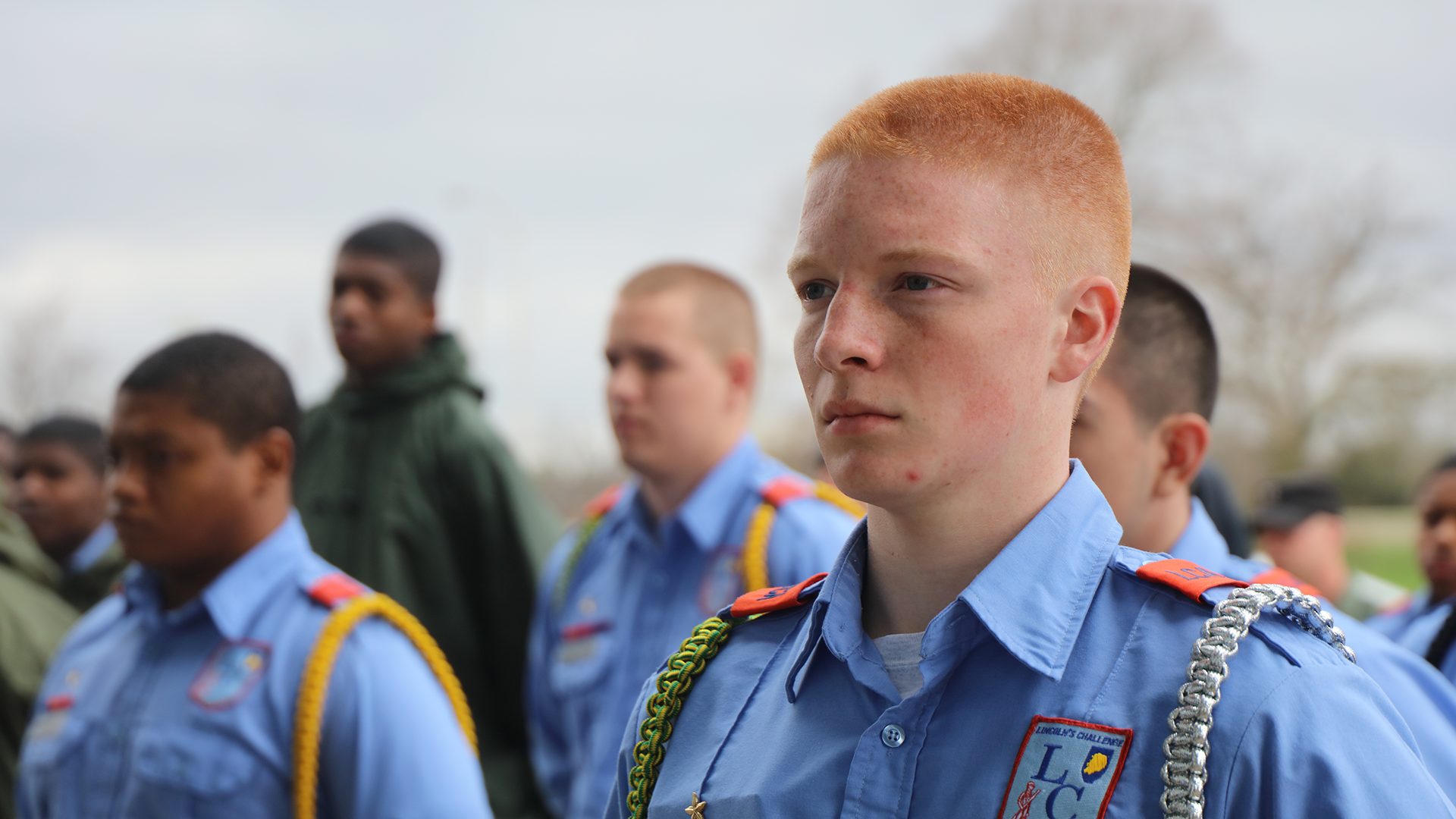 Male cadets in light blue uniforms with aiguillettes standing in formation outdoors