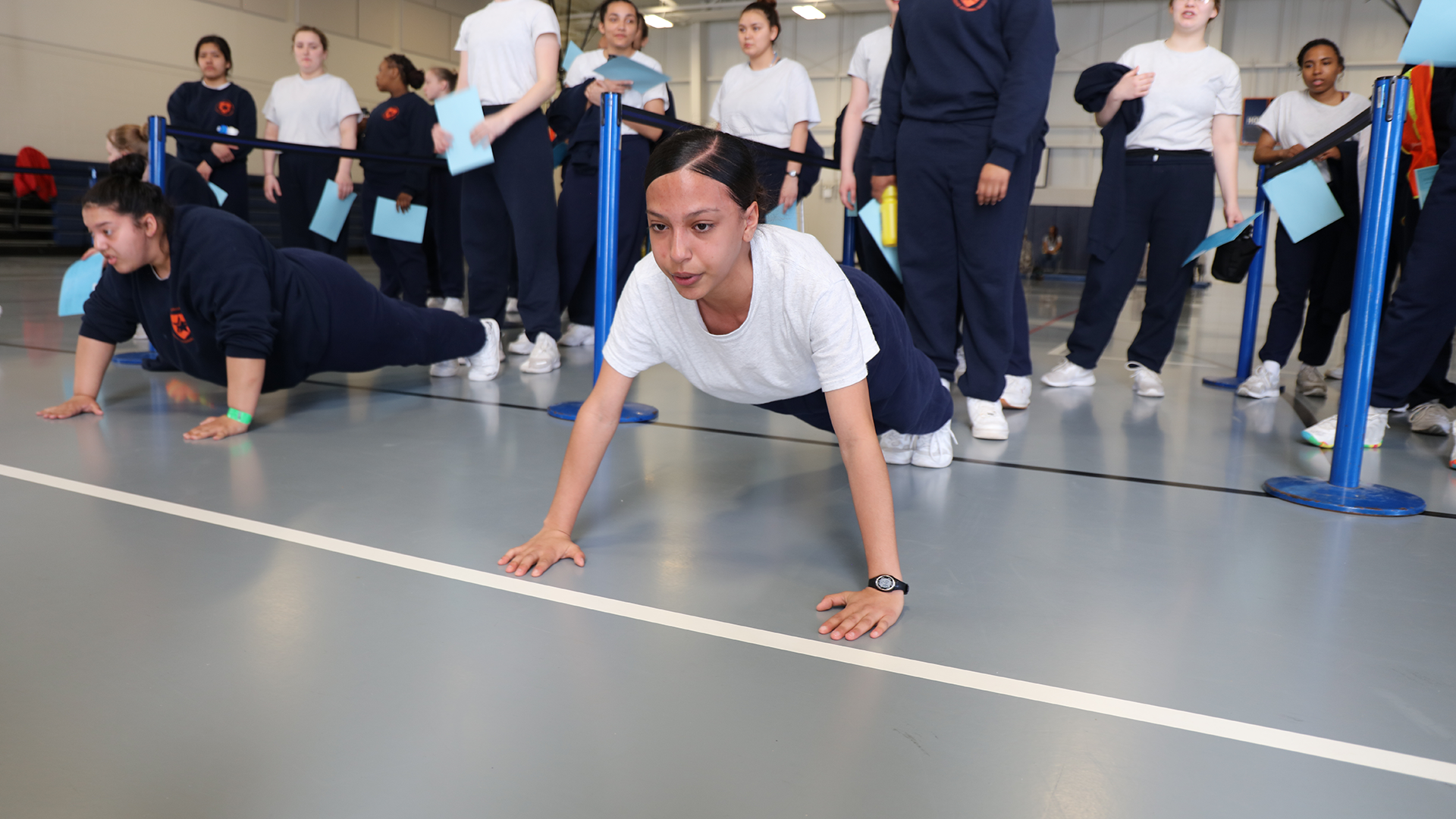 Female cadets doing push-ups on gym floor during physical fitness testing in white shirts and navy pants