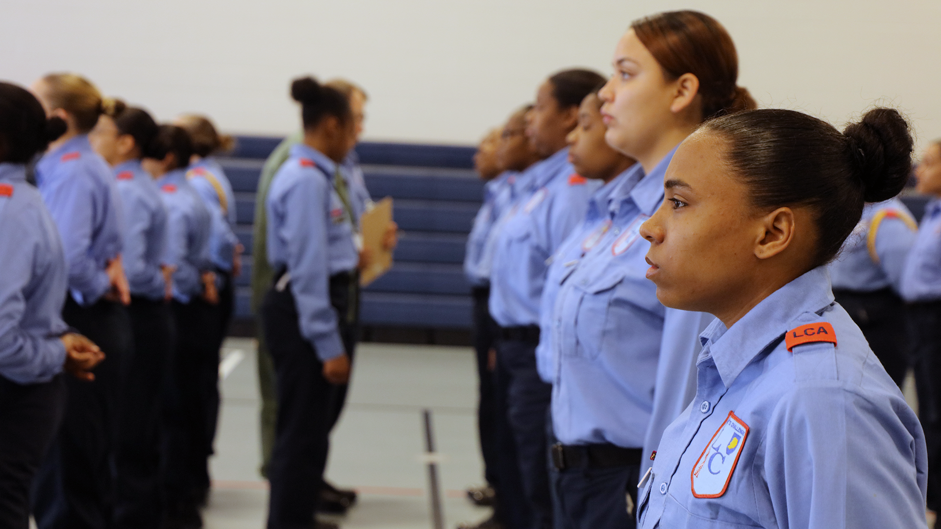 Female cadets in light blue uniforms standing at attention in gymnasium during indoor formation