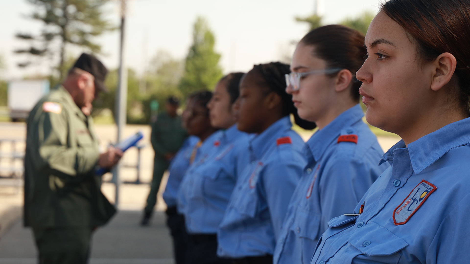 Female cadets in light blue uniforms at attention while drill instructor reviews formation outdoors
