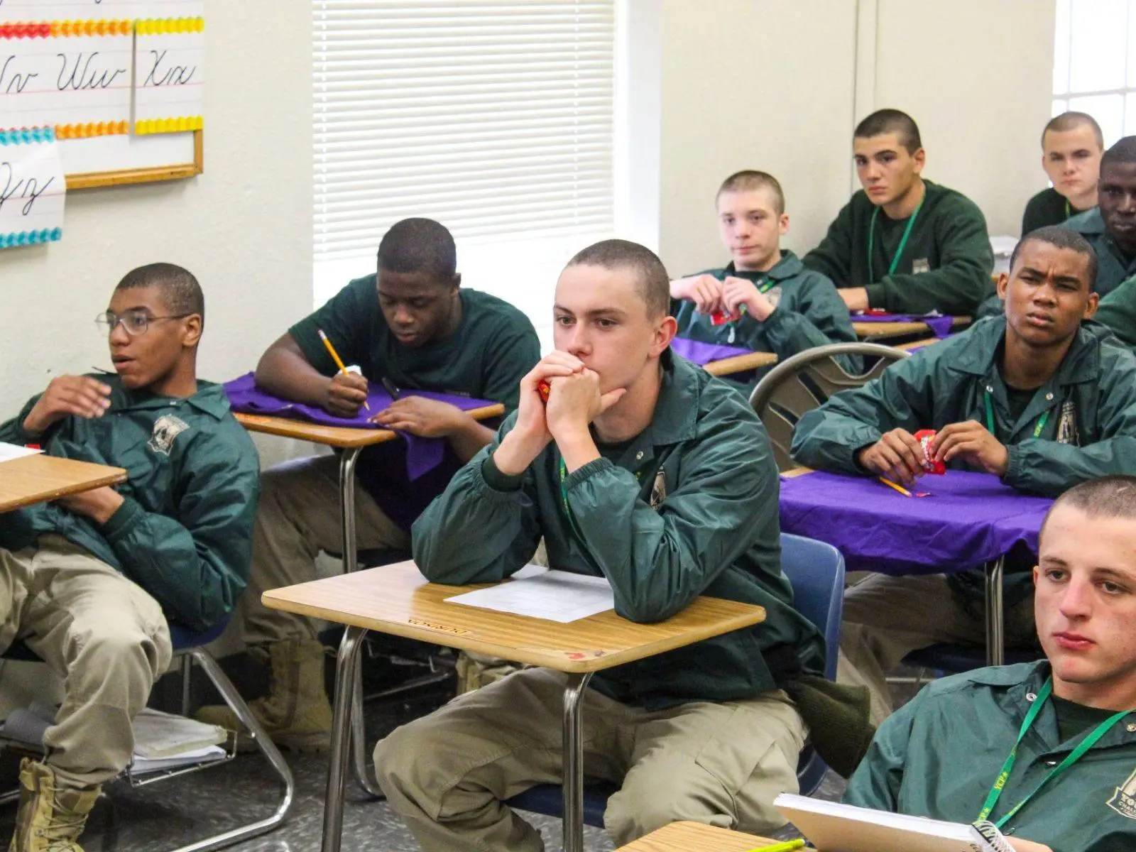 Male cadets with military haircuts in green jackets seated at desks during classroom instruction
