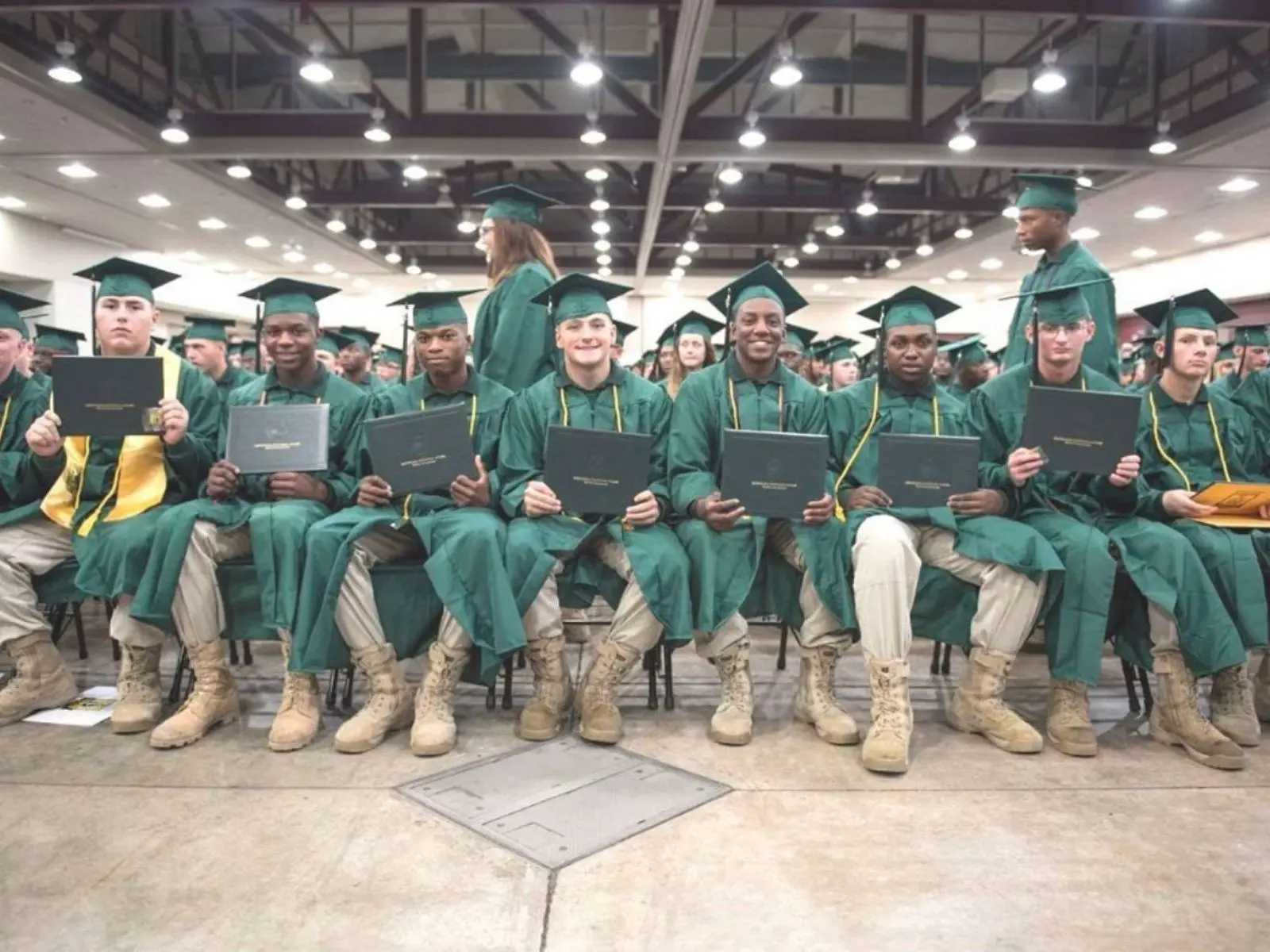 Graduates in green caps and gowns with combat boots holding diplomas at indoor graduation ceremony