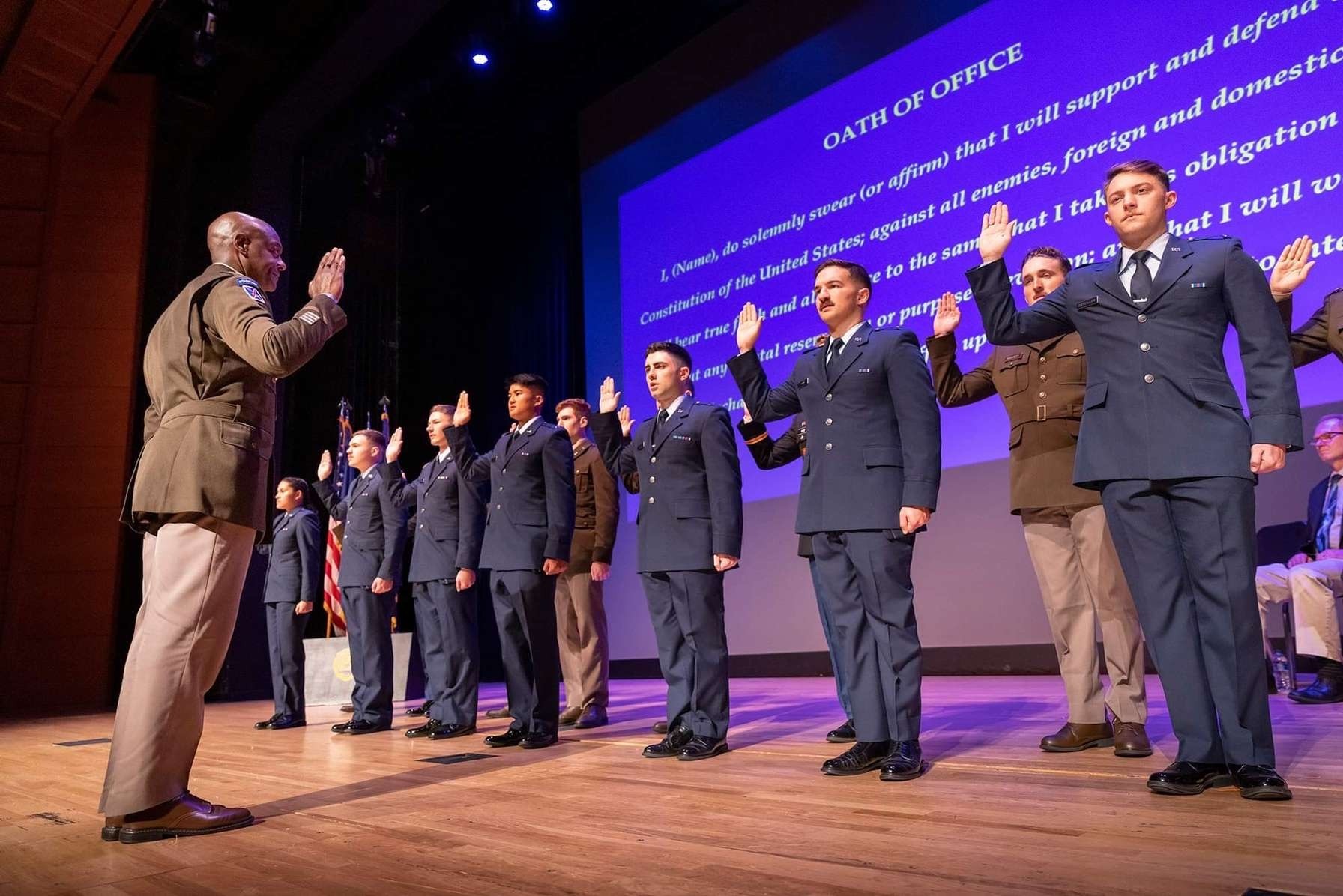 ROTC cadets in dress uniforms taking Oath of Office on stage with projected text behind them