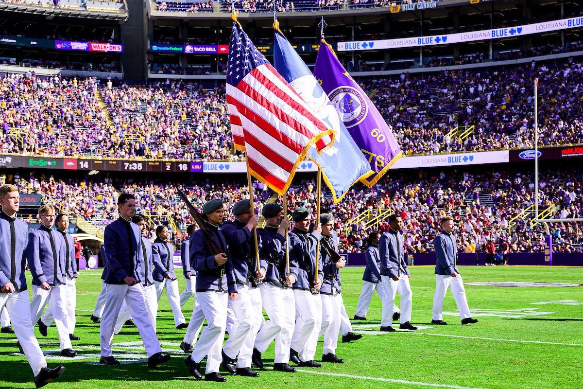Cadets in gray and white uniforms marching with color guard carrying US and LSU flags at Tiger Stadium