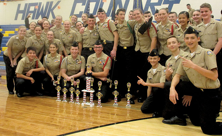NJROTC cadets in khaki uniforms posing with drill competition trophies in gymnasium
