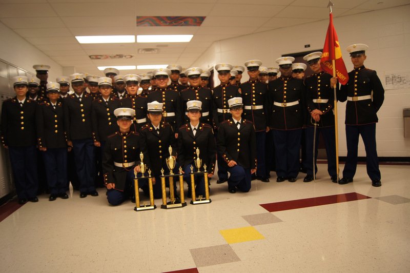 MCJROTC drill team in Marine dress blue uniforms with white covers posing with trophies and guidon