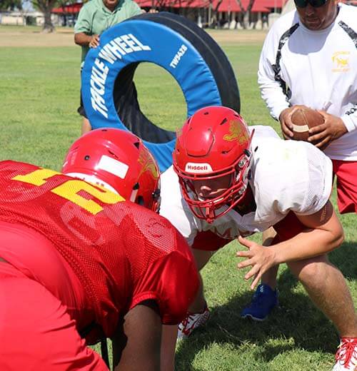 Football players in scarlet and gold helmets practicing tackling drills with coach holding football
