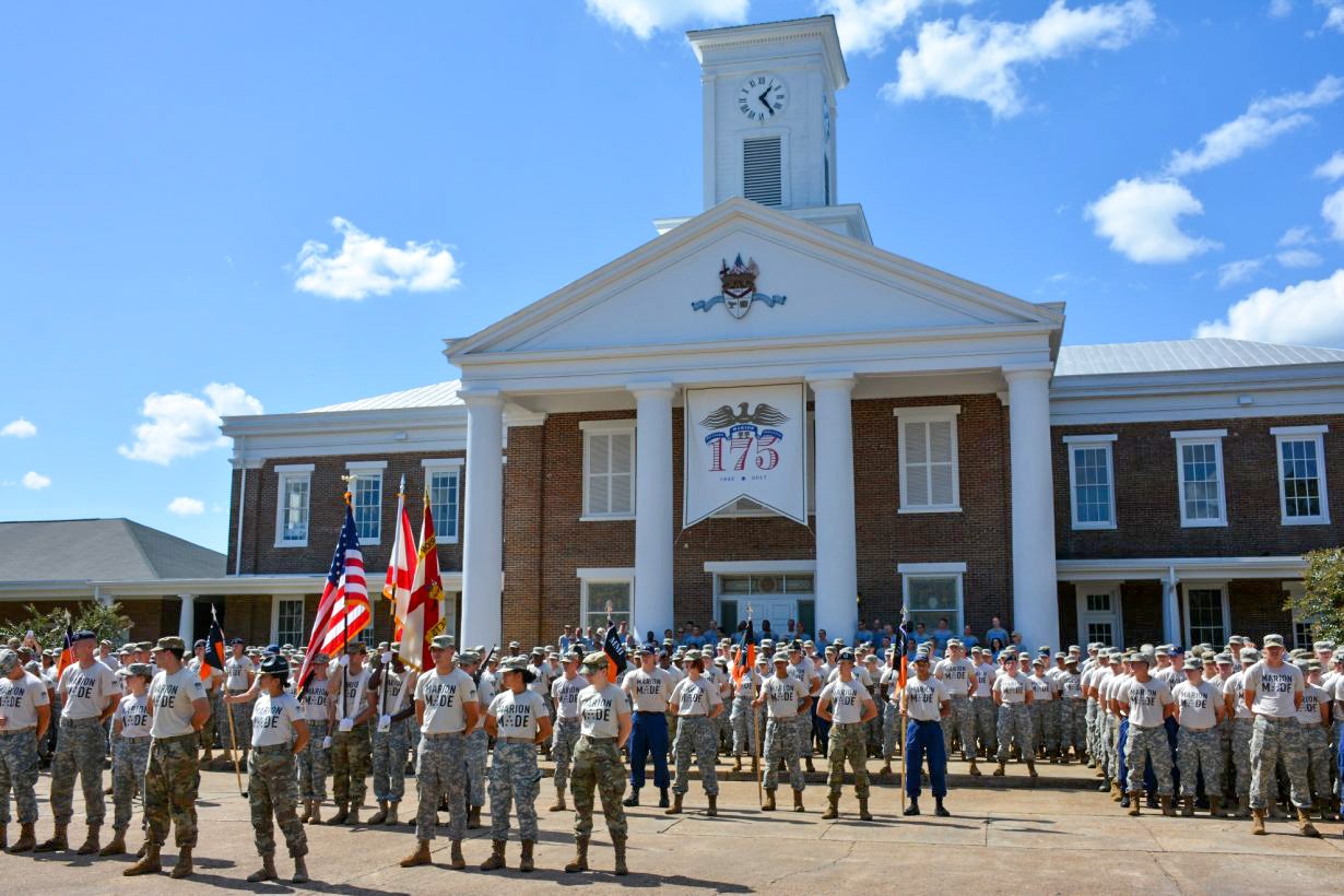 Cadets in formation wearing Marion Made t-shirts and camo pants with color guard flags in front of main hall clock tower