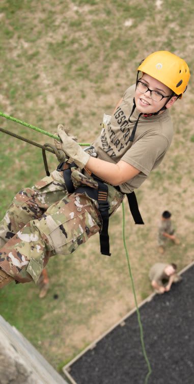 Cadet in camouflage and yellow helmet rappelling down building during training exercise