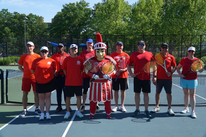 Tennis team in red shirts posing with Warrior mascot in Roman centurion costume on outdoor court