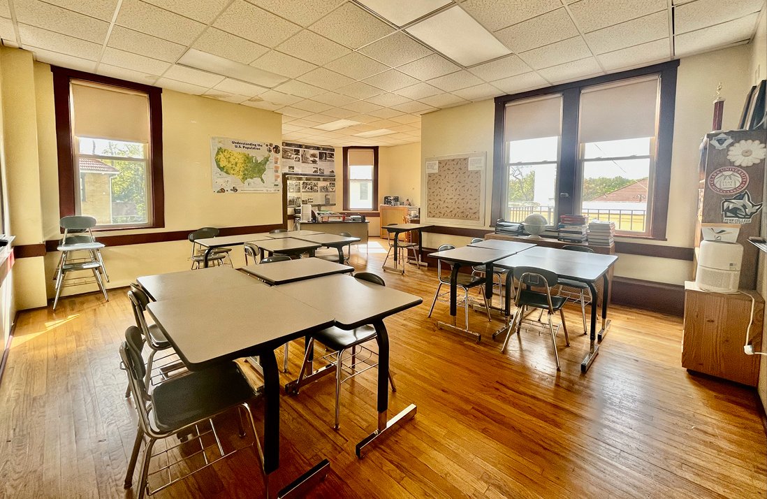 Sunlit classroom with hardwood floors, grouped desks, US map poster, large windows overlooking campus grounds