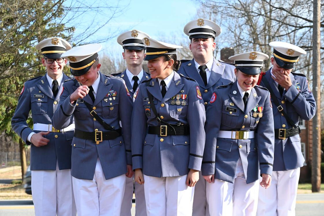 Cadets in gray dress uniforms with white caps and medals laughing together outdoors on sunny day