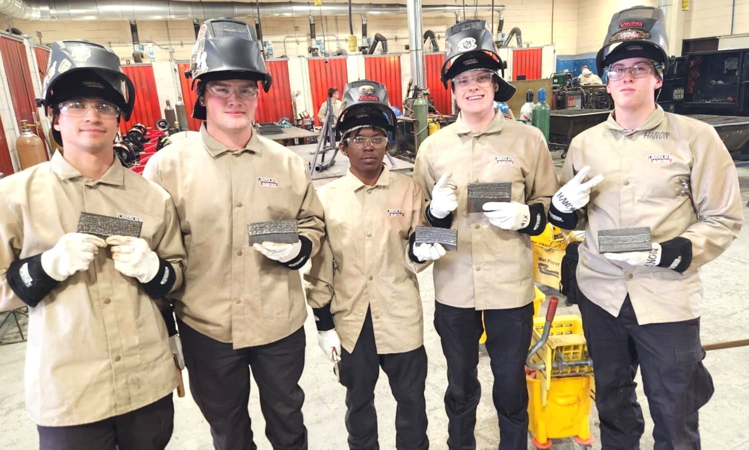 Five welding students in tan jackets and helmets proudly displaying their completed welding projects