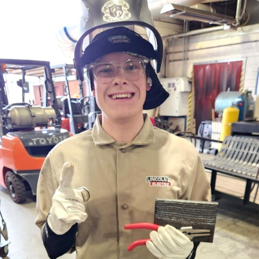 Smiling welding student in tan jacket and safety helmet giving thumbs up while holding welded metal piece