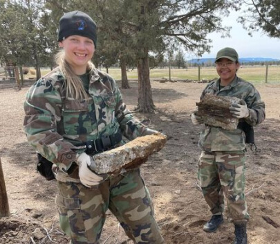 Two female cadets in camouflage uniforms carrying firewood during outdoor community service project