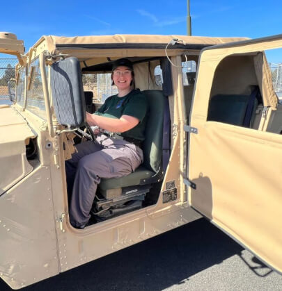 Smiling cadet in green cap and uniform seated in tan military Humvee vehicle on campus