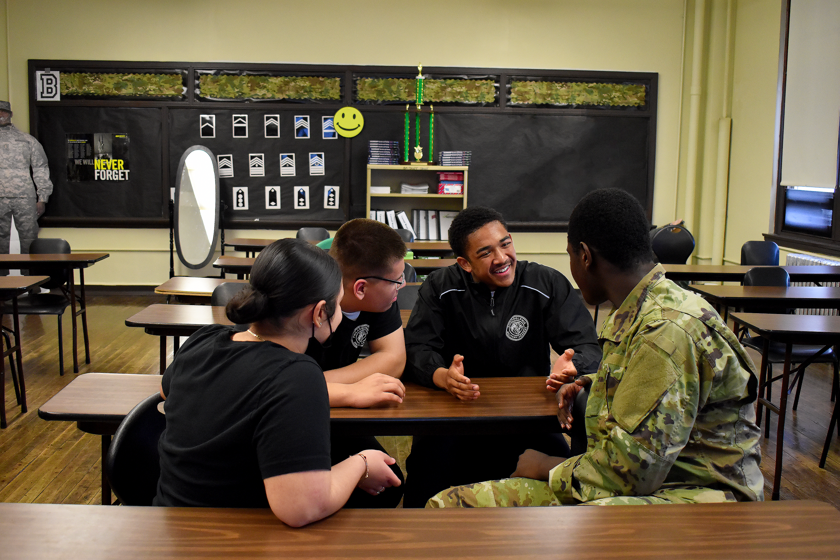 Cadets in JROTC classroom with bulletin board showing rank insignia and Never Forget display
