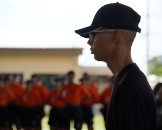 Cadet in black uniform and cap standing at attention in profile with cadets in orange shirts in background