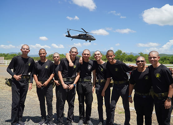 Group of cadets in black PT uniforms posing on airfield with Black Hawk helicopter flying overhead at Fort Allen