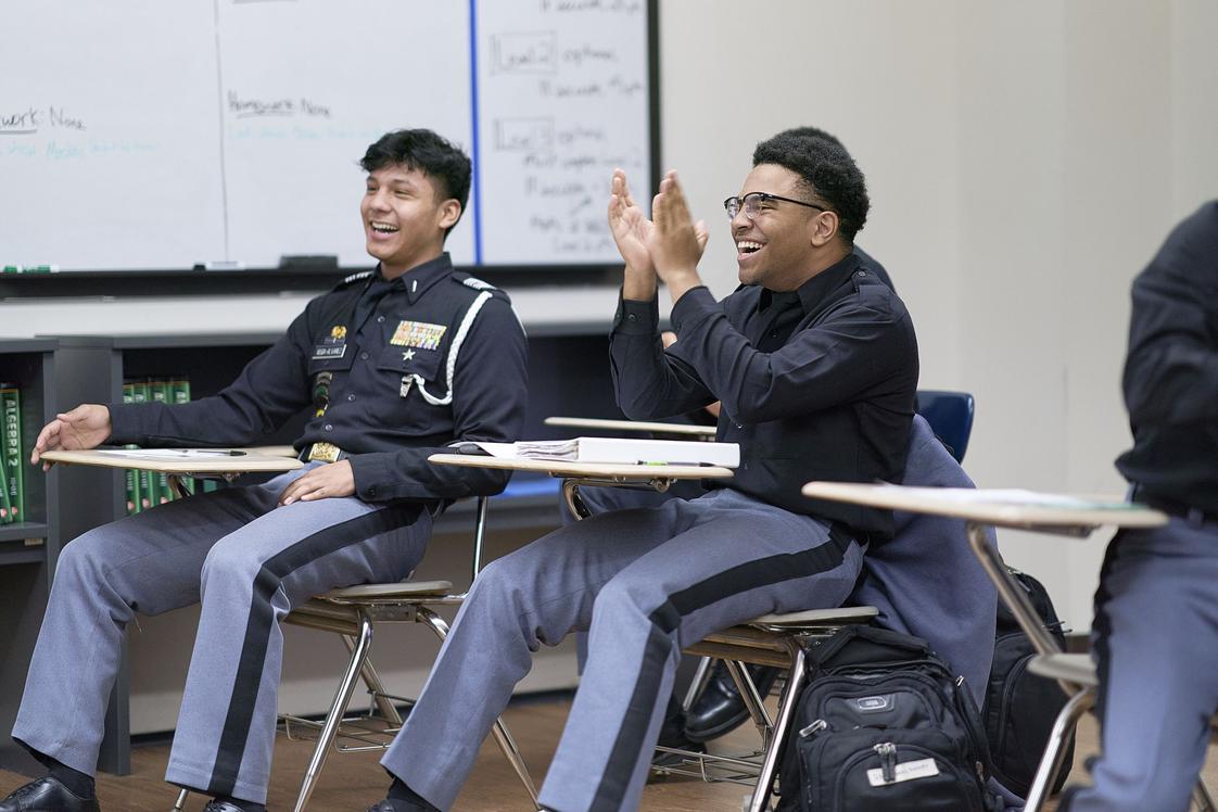 Cadets in dark dress uniforms laughing and clapping at desks during classroom discussion