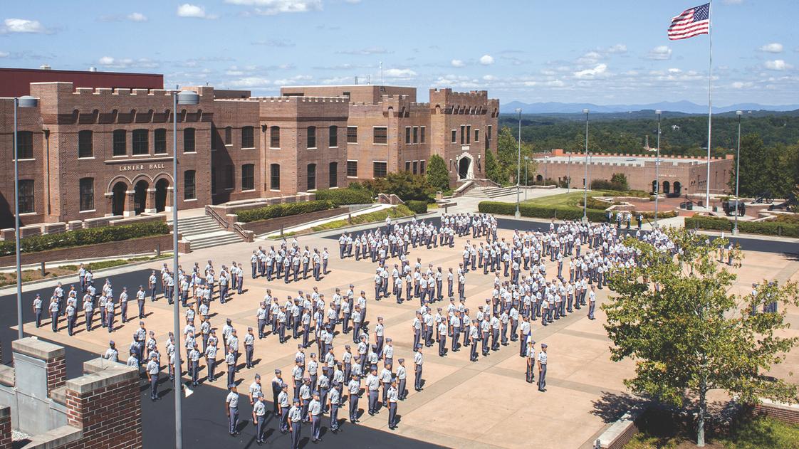 Cadets in gray uniforms standing in formation on parade grounds with Lanier Hall and American flag