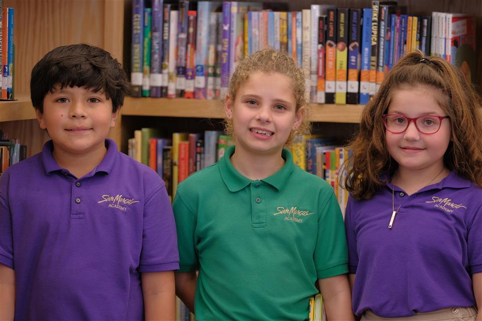 Three elementary students in purple and green SMA polo shirts smiling in school library