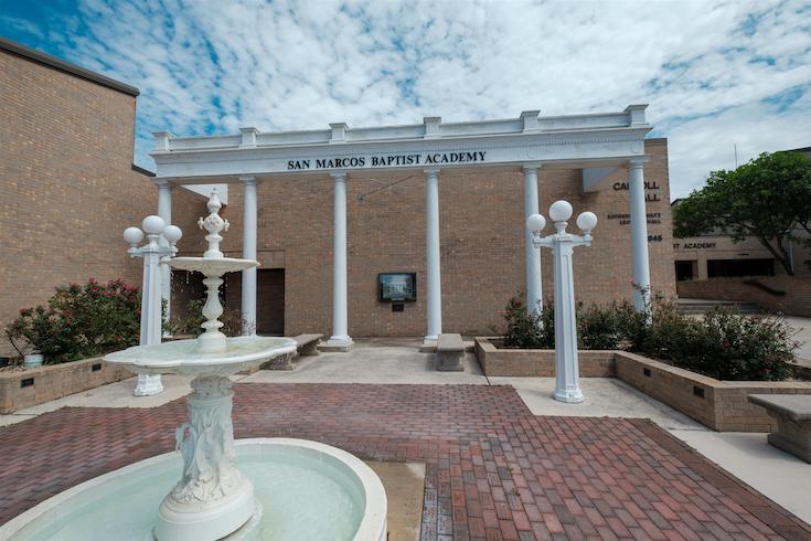 White fountain and columned pergola entrance with San Marcos Baptist Academy sign on brick plaza
